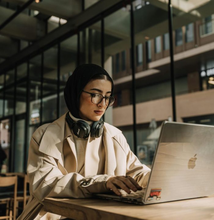 A focused woman wearing a headscarf works on her laptop in a stylish urban cafe.