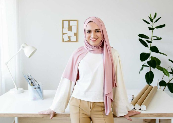 A smiling woman in a pink hijab stands confidently by a desk in a modern well-lit office setting.