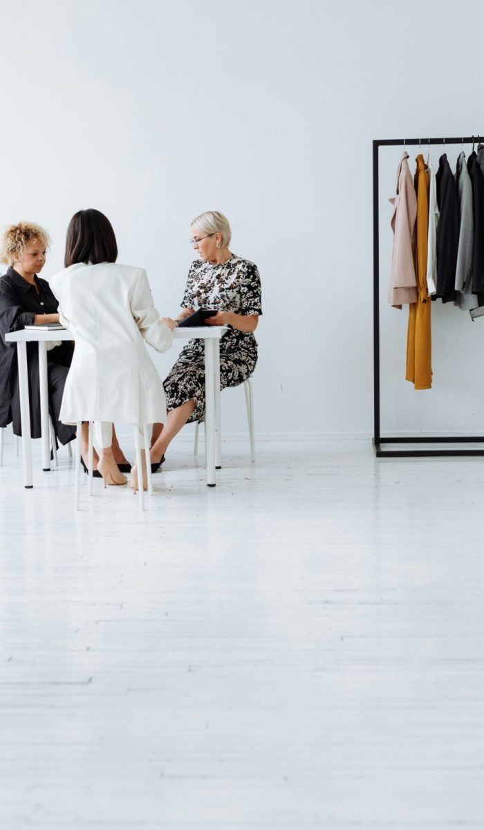 Three women collaborating in a modern, minimalistic office setting with fashionable clothing on display.