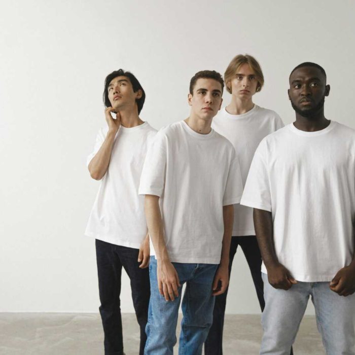 A diverse group of young men stand together indoors, each wearing a casual white t-shirt.